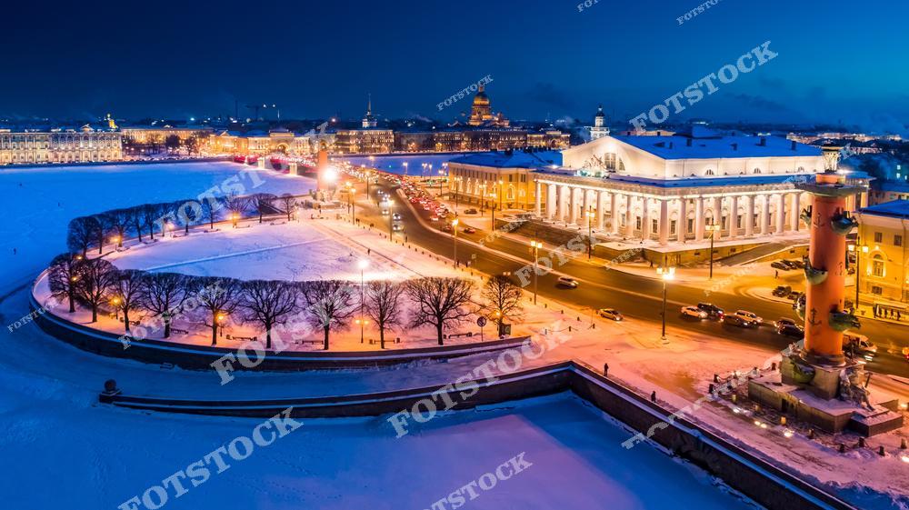 На фотографии изображена Стрелка Васильевского острова зимой, с центральными элементами — Ростральными колоннами и зданием Биржи. Снежный покров и вечернее освещение подчеркивают величественный облик исторического центра Санкт-Петербурга. Вдалеке виден Исаакиевский собор.